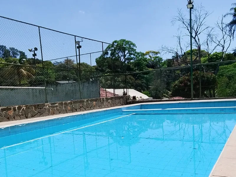 Piscina de azulejos azuis com borda de pedra clara, cercada por alambrado e árvores
